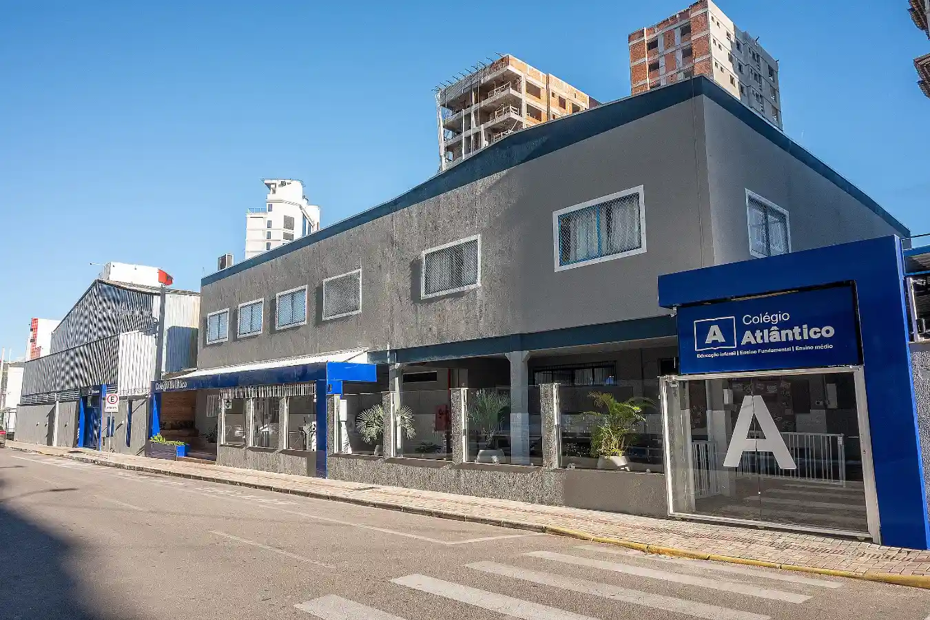 Fachada principal do Colégio Atlântico em Itapema, com estrutura moderna e fachada cinza e azul, representando as etapas da Educação Infantil, Ensino Fundamental e Médio.